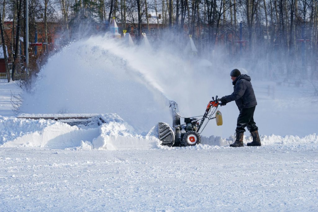 Mann in warmer Kleidung geht mit einer Schneefräse und reinigt seinen Garten in der Stadt.